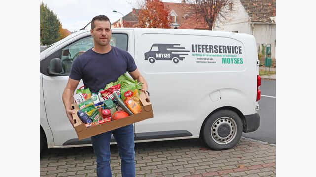 Manfred Moyses, Obmann des burgenländischen Lebensmittelhandels, mit einer Kiste Lebensmittel vor seinem weißen Lieferwagen.