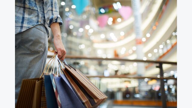 Nahaufnahme auf eine Person von hinten, die in einem hell erleuchteten Shoppingcenter mehrere Einkaufstaschen trägt