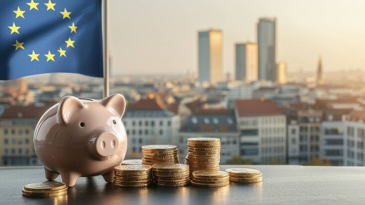 Ein Sparschwein und Münzen vor dem Hintergrund einer EU-Flagge und weiter hinten eine Stadt