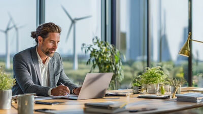Mann am Laptop vor großen Fenstern mit Windrädern im Hintergrund