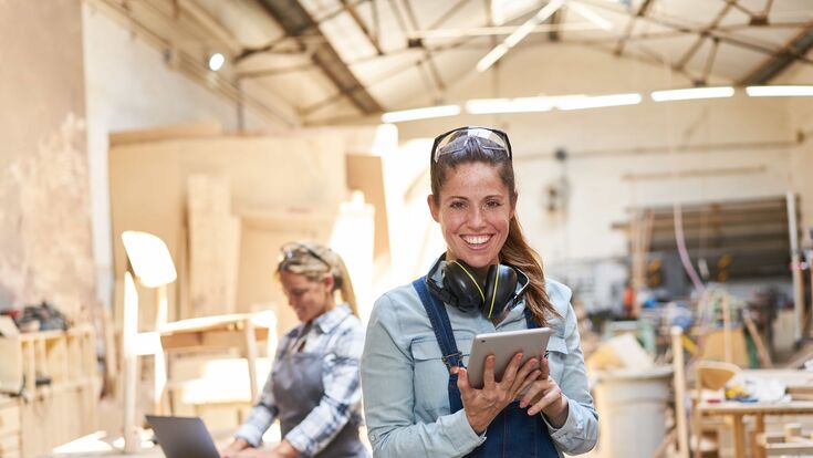 Eine lächelnde, junge Tischlerin in der Werkstatt, Tablet in den Händen, Schutzbrille hochgeschoben