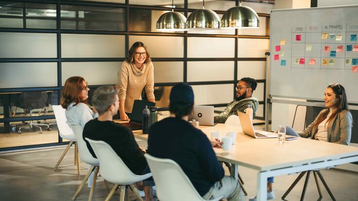 Sechs junge Männer und Frauen sitzen in lockerer Atmosphäre um einen Meeting-Tisch, eine der Frauen steht.