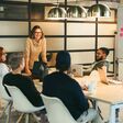 Sechs junge Männer und Frauen sitzen in lockerer Atmosphäre um einen Meeting-Tisch, eine der Frauen steht.
