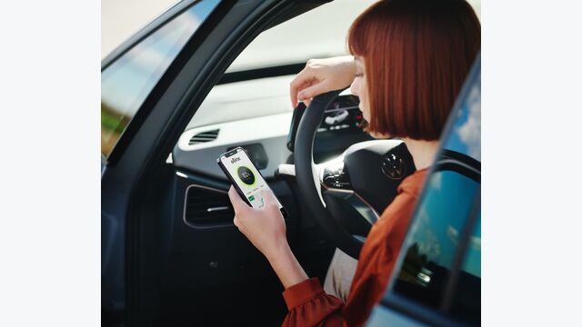 Eine Frau sitzt in ihrem Auto hinter dem Lenkrad und blickt auf ihre E-Lade-App am Smartphone.