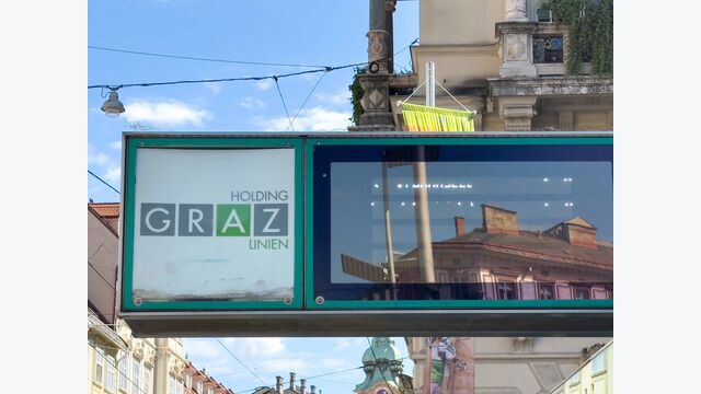 Öffi-Anzeigetafel der Holding Graz Linien, in der sich ein großes Gebäude spiegelt.