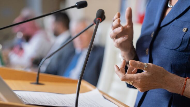 Nahaufnahme einer Frau in Business-Kleidung an einem Podium eine Rede haltend, Kopf nicht sichtbar.