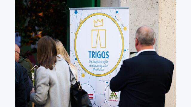 Ein großer Banner auf der Trigos-Verleihung 2025 in Wien, daneben Gäste.