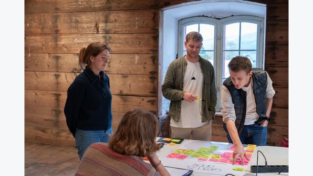 Vier junge Menschen stehen und sitzen bei der Renowave-Sanierungsmesse um einen Tisch mit Poster und Klebenotizen vor sich.