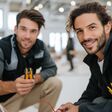 zwei lächelnde junge Männer hocken vor aus der Wand ragenden Kablen, Abisolierzangen in Händen