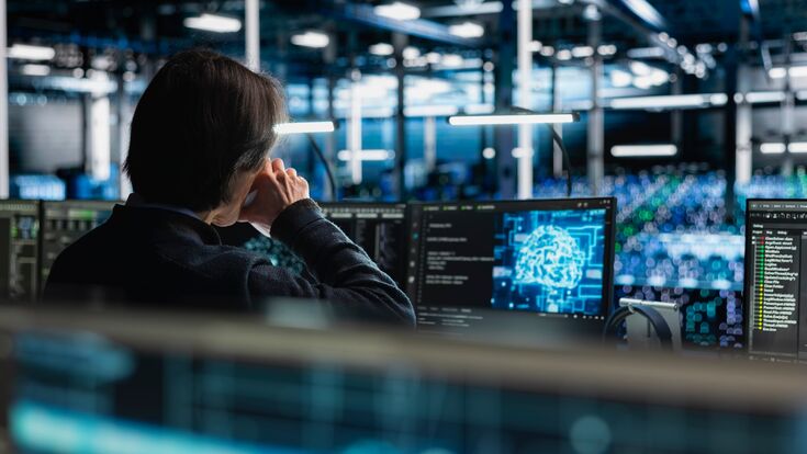 Rückenansicht auf einen Menschen in großem dunklen Raum mit vielen Computern, der Bildschirm vor ihm zeigt leuchtendes neuronales netzwerk