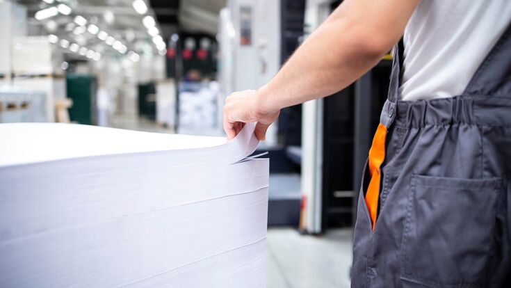 Industriearbeiter (von hinten, Ausschnitt) berührt einen hohen Stapel großer weißer Papierbögen, Fabriksumgebung