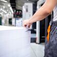 Industriearbeiter (von hinten, Ausschnitt) berührt einen hohen Stapel großer weißer Papierbögen, Fabriksumgebung