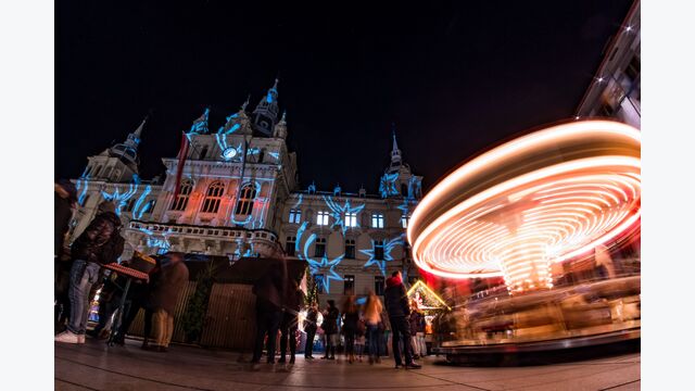 Grazer Christkindlmarkt im Dunkeln mit BesucherInnen und hell leuchtendem, sich schnell drehenden Karussell.