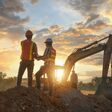zwei bauleiter mit sicherheitshelmen und warnwesten von hinten auf dem erdhügel eine baustelle, bagger und sonnenuntergang im hintergrund.
