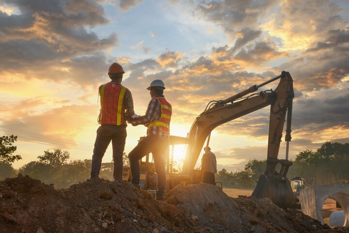 zwei bauleiter mit sicherheitshelmen und warnwesten von hinten auf dem erdhügel eine baustelle, bagger und sonnenuntergang im hintergrund.