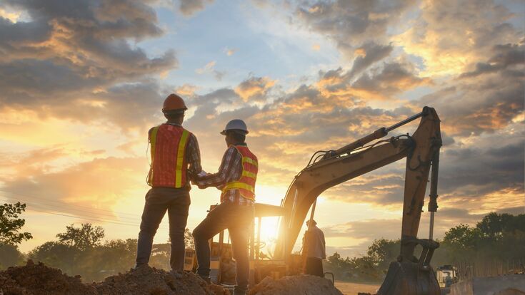 zwei bauleiter mit sicherheitshelmen und warnwesten von hinten auf dem erdhügel eine baustelle, bagger und sonnenuntergang im hintergrund.