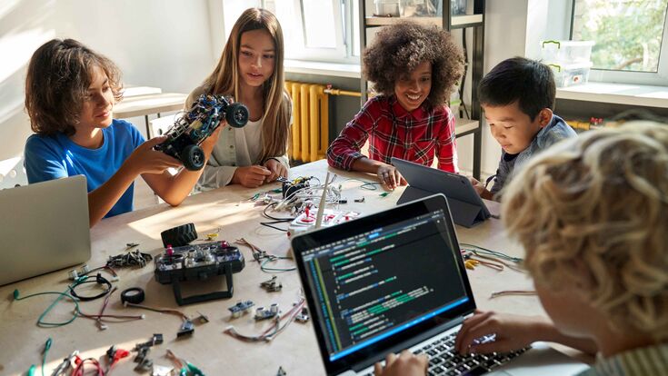 Kinder sitzen um einen Tisch, bauen kleine Autos und programmieren am Laptop