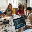 Kinder sitzen um einen Tisch, bauen kleine Autos und programmieren am Laptop