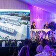 Leobersdorfer maschinenfabrik-CEO Günther Ognar auf der bühne der jubiläumsfeier mit moderatorin marie-claire zimmerman, im hintergrund ein großes bild der fabrik