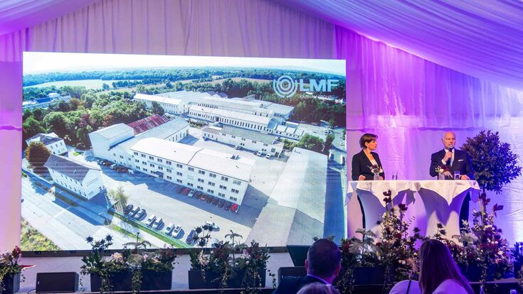 Leobersdorfer maschinenfabrik-CEO Günther Ognar auf der bühne der jubiläumsfeier mit moderatorin marie-claire zimmerman, im hintergrund ein großes bild der fabrik