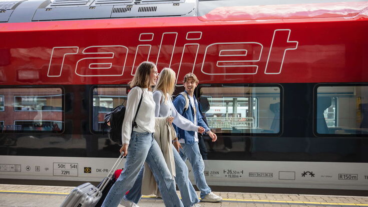 drei fröhliche Jugendliche ziehen kleine koffer am Bahnsteig vor einem öbb Railjet