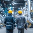 Rückenansicht auf zwei Arbeiter mit Schutzhelmen in einer großen Halle mit einem riesigen Rohrsystem in der Energieindustrie