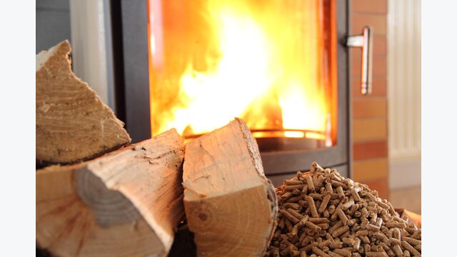 Holzscheite und Pellets liegen vor einem Ofen mit Glastür, in dem ein helles Feuer brennt