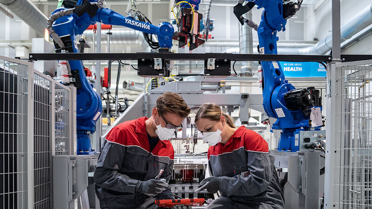 Ein männlicher und ein weiblicher Lehrling mit Schutzmasken beugen sich über ein Tablet mit großen Roboterarmen im Hintergrund