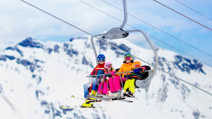 Vierköpfige Familie in voller Skiausrüstung auf einem Sessellift, im Hintergrund verschneiter Berg