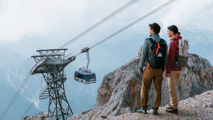 Ein Pärchen mit Wanderrucksäcken steht auf einem Bergvorsprung und blickt auf die Tiroler Zugspitzbahn