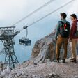 Ein Pärchen mit Wanderrucksäcken steht auf einem Bergvorsprung und blickt auf die Tiroler Zugspitzbahn