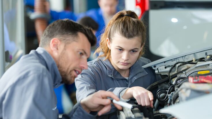 junge frau (weiblicher lehrling) wird von einem mann in der automechanik angeleitet