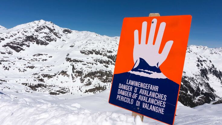 ein oranges schild vor einem verschneiten berg zeigt eine warnende hand und warnt vor lawinengefahr