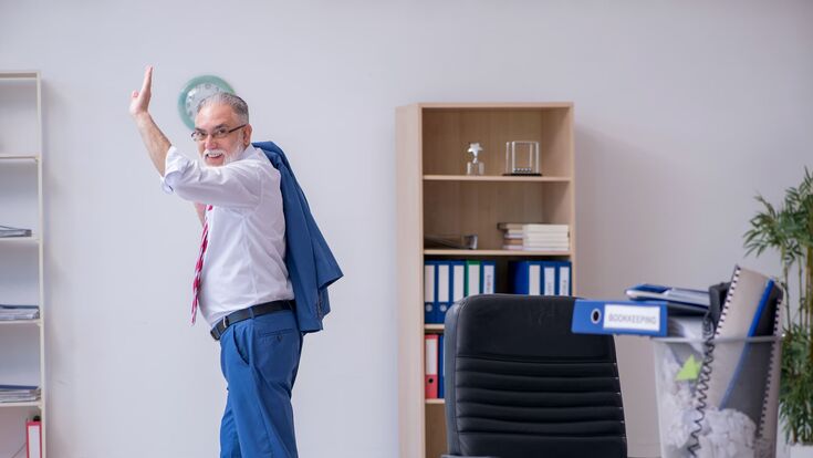 alter mann in anzug winkt glücklich, während er im büro schreibtisch und papierkram hinter sich lässt
