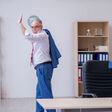 alter mann in anzug winkt glücklich, während er im büro schreibtisch und papierkram hinter sich lässt