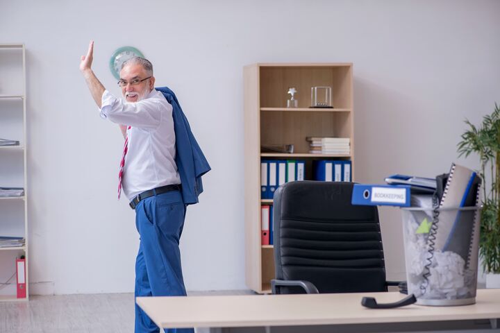 alter mann in anzug winkt glücklich, während er im büro schreibtisch und papierkram hinter sich lässt