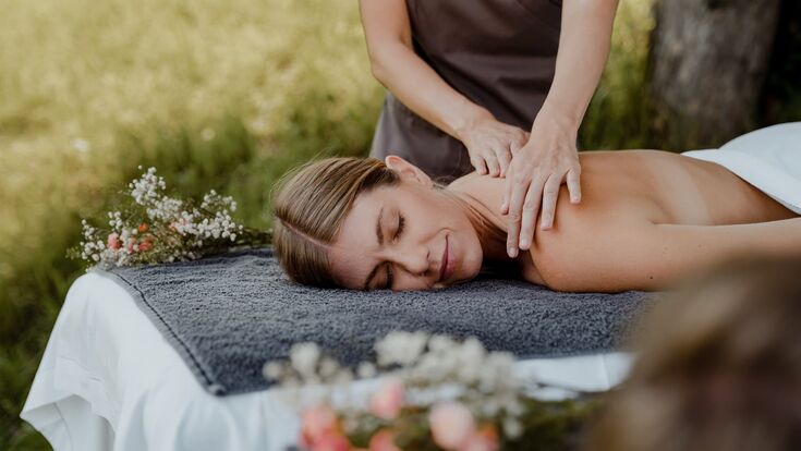 junge attraktive frau wird auf massagebank massiert, daneben blumen, im hintergrund wiese