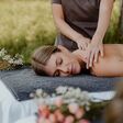junge attraktive frau wird auf massagebank massiert, daneben blumen, im hintergrund wiese