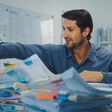 junger unternehmer sucht an einem schreibtisch in hohen stapeln geordnetem papier