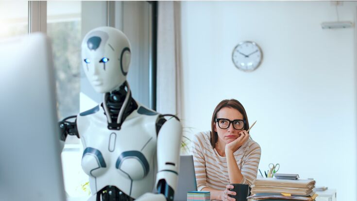 eine junge frau an einem schreibtisch im büro blickt skeptisch auf einen humanoiden roboter am schreibtisch vor ihr bei der computerarbeit