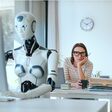 eine junge frau an einem schreibtisch im büro blickt skeptisch auf einen humanoiden roboter am schreibtisch vor ihr bei der computerarbeit