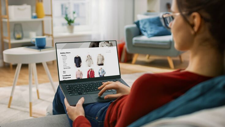 eine frau sitzt auf dem sofa und sieht sich auf dem laptop damenmode auf einem onlinehandel-portal an