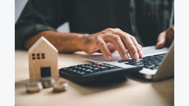 händes eines mannes tippen auf laptop, daneben taschenrechner, kleines holzhaus und münzen: symbolisch für hauskauf und kreditberechnung