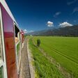 blick aus pinzgauer lokalbahn auf grüne wiese unter blauem himmel, teil der bahn sichtbar